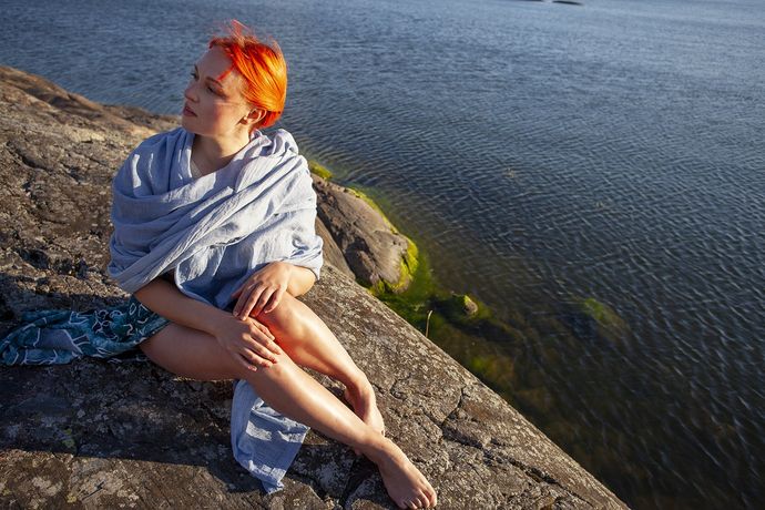 A person with orange hair sits wrapped in a light blue shawl on a rocky shore, looking out over a calm body of water on a sunny day.