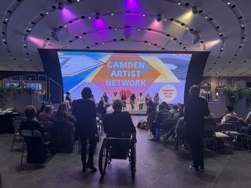 A diverse audience attends the Camden Artist Network Late Summer Social 2025, featuring a panel discussion under a large, colourful screen.