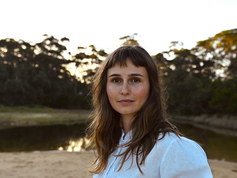 Person with fringe and long brown hair looks to camera with lake and trees behind
