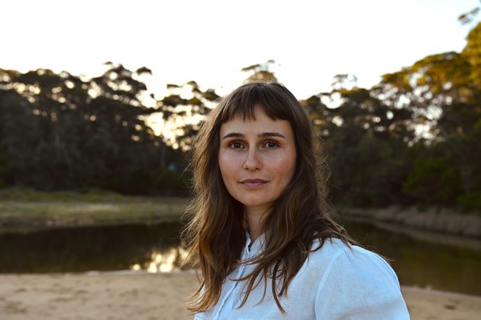 Person with fringe and long brown hair looks to camera with lake and trees behind