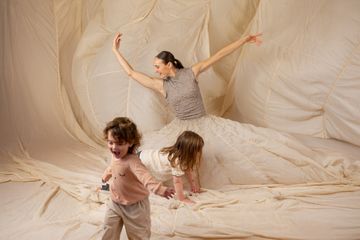 A woman in a flowing dress poses gracefully while two children play in front of her against a soft, draped fabric backdrop.