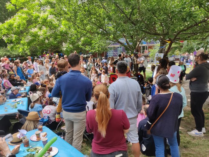 Lots of people of different ages - many children - gather under 3 leafy green trees outdoors in park, wearing colourful summer clothed, standing near picnic tables