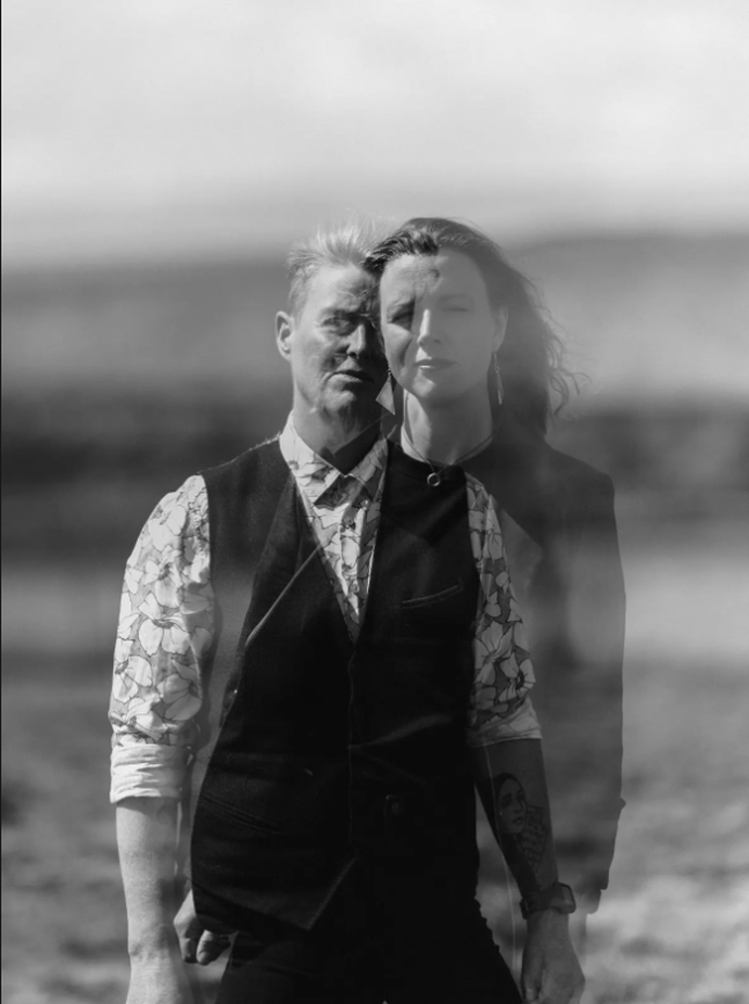 A black and white double exposure of two people standing outdoors, blending their images against a blurred natural background.