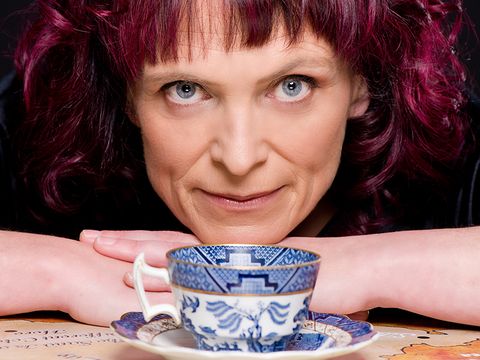 Person with curly purple hair resting arms on a table, looking forward, with a blue and white teacup and saucer in the foreground.