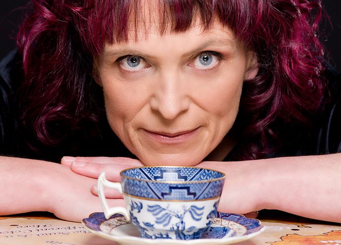 Person with curly purple hair resting arms on a table, looking forward, with a blue and white teacup and saucer in the foreground.