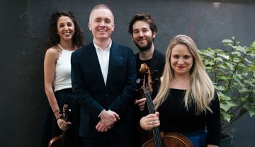 Four people standing together indoors, three holding string instruments including cellos, with a potted plant in the background.