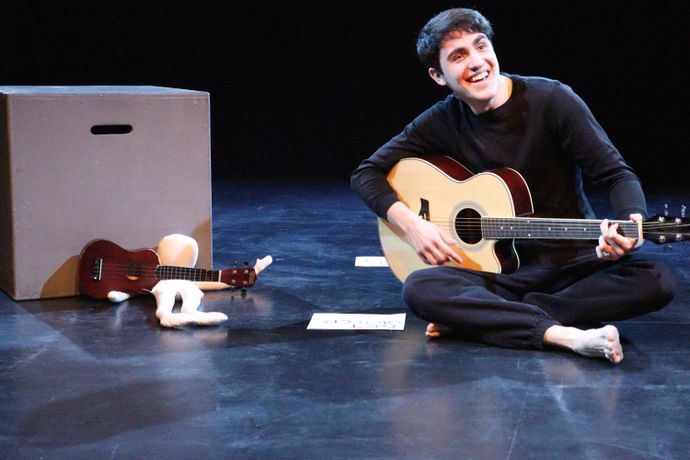 Musician sits on the floor with guitar in hand accompanies by a plushie who is holding a musical instrument. A note is also displayed on the floor.