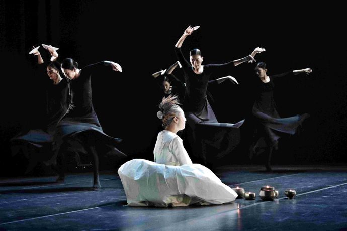 A seated performer in white with hair styled up; five dancers in black move gracefully on a dark stage.