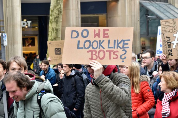 People at a protest hold a sign reading "DO WE LOOK LIKE BOTS?" in a busy urban area with shops in the background.