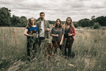 Four musicians standing in a grassy field, each holding a string instrument, with trees and cloudy sky in the background.