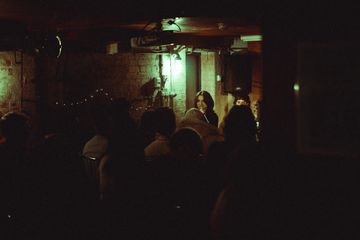 Dimly lit underground music venue with a singer on stage, brick walls, string lights, and an audience seated in shadows.