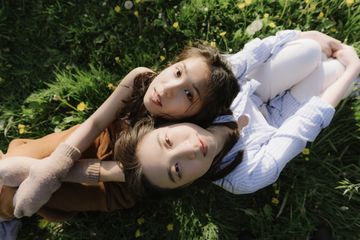 Two people sitting on grass, looking up. One wears a brown outfit, the other in white with a striped shirt. Yellow flowers scattered around.