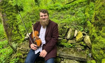 Will Allen sits with a violin on a mossy stone bench in a lush, green forest setting.