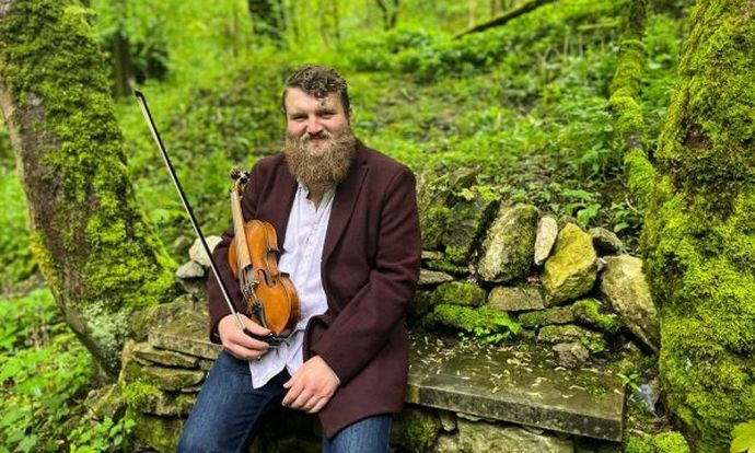 Will Allen sits with a violin on a mossy stone bench in a lush, green forest setting.