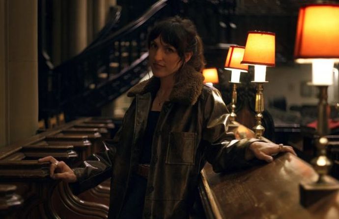 Person wearing a dark leather coat with a fur collar, standing in a dimly lit interior beside wooden pews and a row of warm-lit lamps.