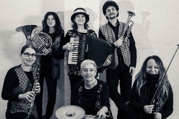 Six musicians pose with instruments, including a trumpet, accordion, clarinet, and French horn, against a plain background.