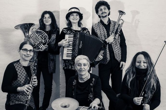 Six musicians pose with instruments, including a trumpet, accordion, clarinet, and French horn, against a plain background.