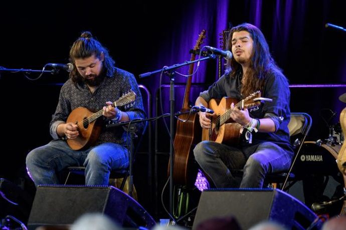 Two musicians seated, playing string instruments on a dimly lit stage, surrounded by microphones and guitars.