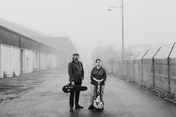 Two people stand on a foggy street, each holding a guitar case, next to industrial buildings and a chain-link fence.