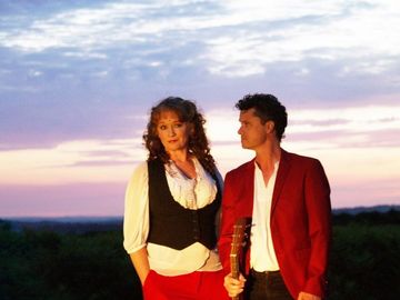 A woman wearing white shirt and black waistcoat with red trousers and long curly hair stands next to man wearing white shirt and red dinner jacket who looks at her. in the background is a sunset dusky sky and fields/forest under