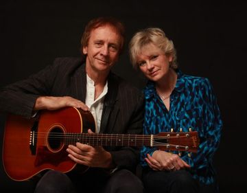 A man and woman sit together, the man holding a guitar. They both smile warmly against a dark background.