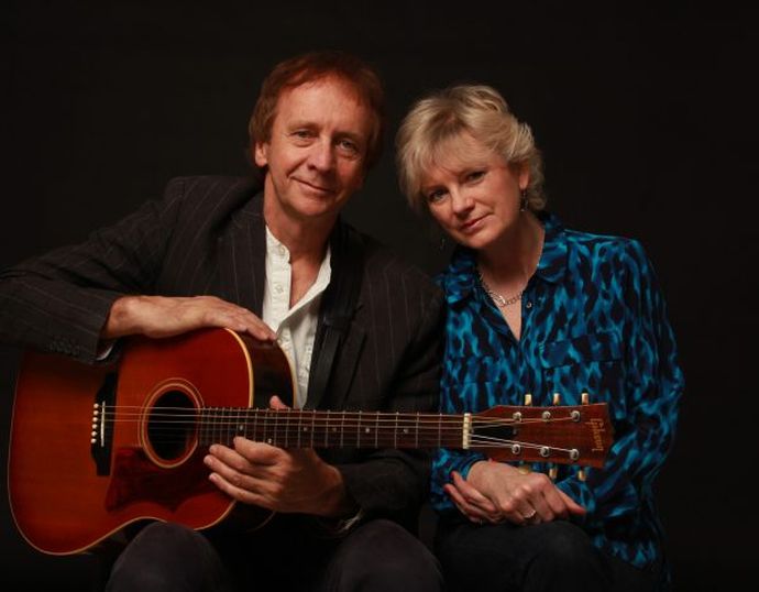 A man and woman sit together, the man holding a guitar. They both smile warmly against a dark background.