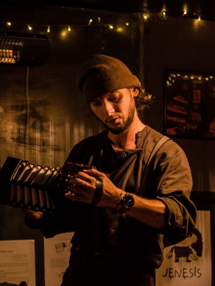 Person wearing a cap and dark clothing playing a concertina in a dimly lit indoor setting. Warm string lights hang above, and framed artwork along with posters are visible on the wall in the background.