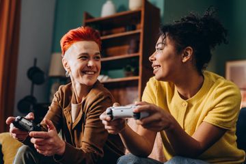 Two people sit indoors holding game controllers and facing each other while engaged in a video game.