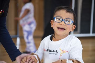 A smiling child wearing blue glasses and a white shirt holds someone's hand indoors. Another child is blurred in the background.
