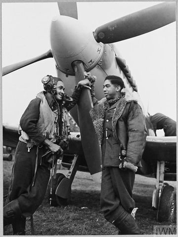 Two pilots in flight gear stand by a propeller of a fighter plane, smiling and talking. One leans casually on the propeller blade.