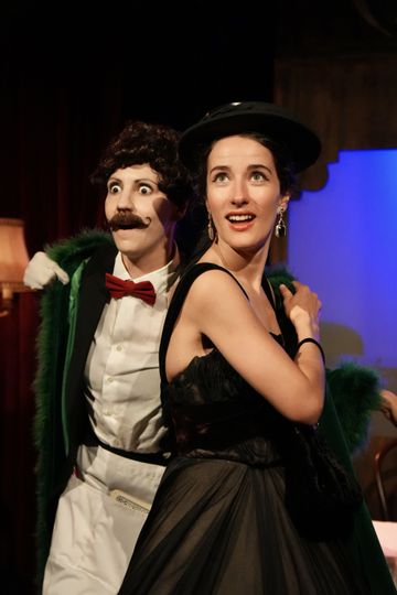 Two performers on stage: one with a painted mustache and tuxedo, the other in an elegant black dress, both with expressive expressions.