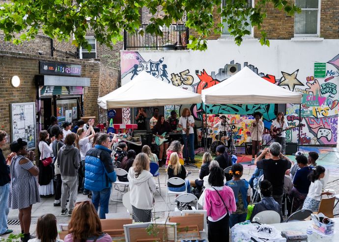 People of all ages stand around in outdoor concrete and brick walled space with colourful graphiti on back wall as musicians play under white gazebos