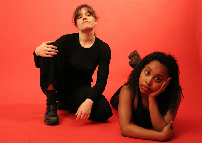 Two people in black outfits pose on a red background. One kneels with hand on knee; the other lies with a hand on their face, looking forward.