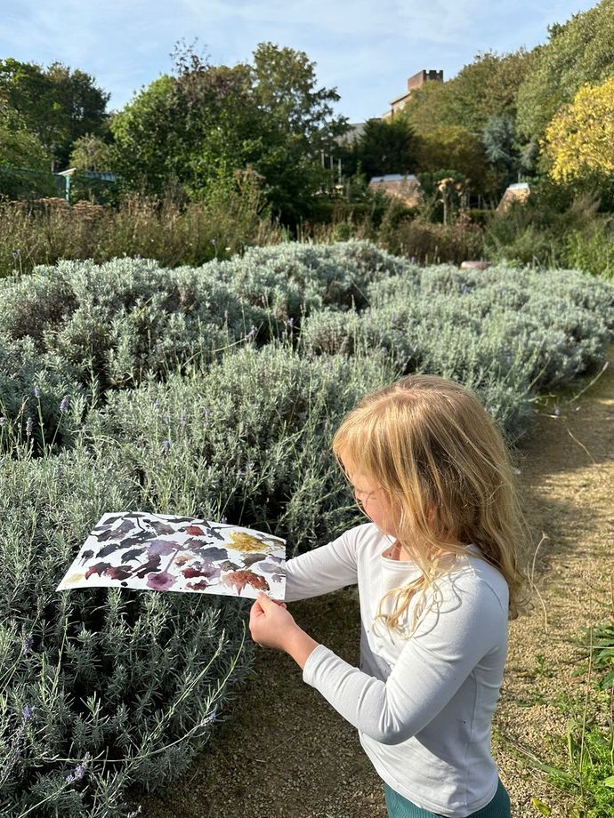 A child stands on a garden path examining a painting held in hand, surrounded by bushes and greenery.