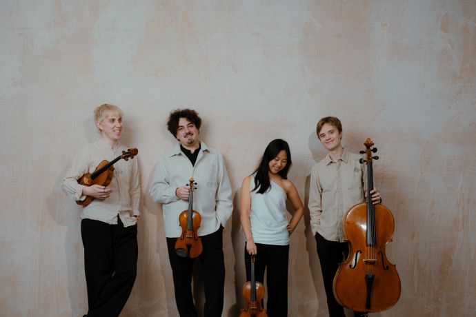 Four musicians standing against a textured wall, holding string instruments, including violins and a cello, casually smiling.