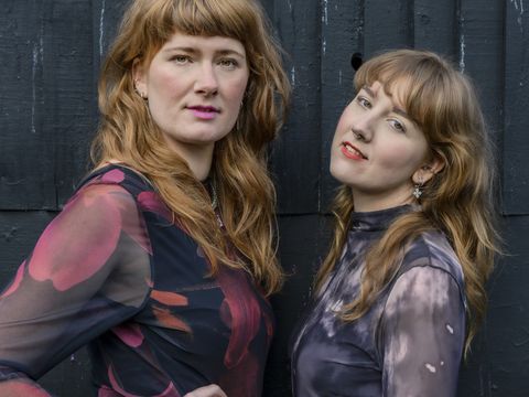 two women with sandy hair and fridnges look to camera with black fence behind them