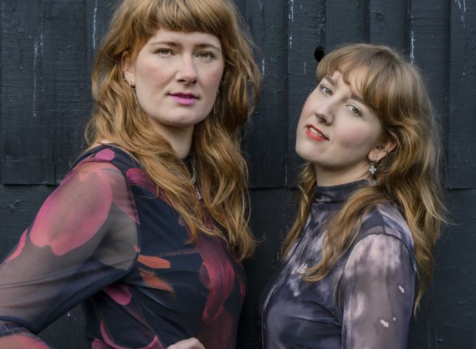 Two people with dark blonde hair standing in front of a black wall
