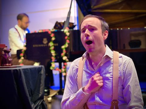 Performer singing in foreground with pianist, stage lighting, decorated piano, and indoor theatre setting.