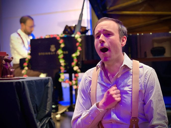 Performer singing in foreground with pianist, stage lighting, decorated piano, and indoor theatre setting.