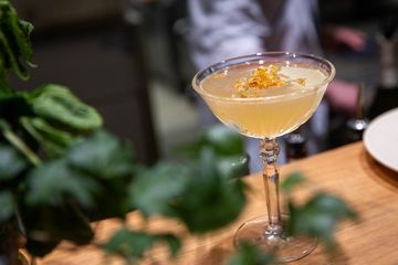 Pale yellow cocktail in a coupe glass garnished with edible flower petals, sitting on a wooden bar top.