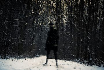 A blurred figure stands on a snowy path surrounded by dark, leafless trees, creating a mysterious and eerie atmosphere.