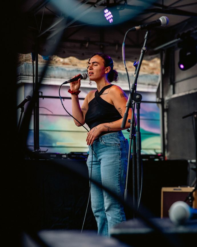 A woman sings passionately on an outdoor stage, holding a microphone. She wears a black top and jeans, with colorful lights in the background.