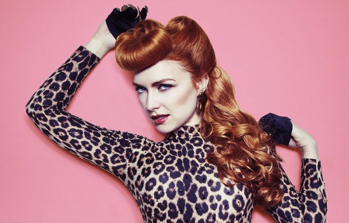 A woman with ginger hair, wearing a leopard print costume, standing in front of a pink background