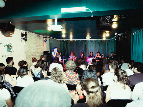 Indoor setting with band on stage and people packed into seats in room with exposed brick and black curtains behind stage