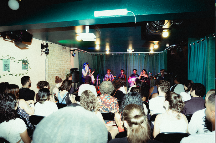 Indoor setting with band on stage and people packed into seats in room with exposed brick and black curtains behind stage