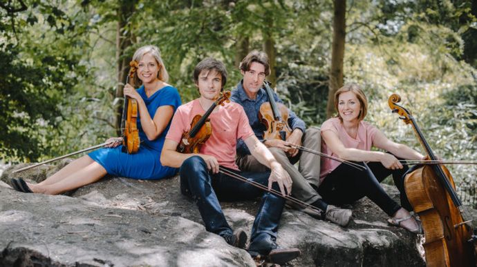 Four people sitting on a rock outdoors, each holding a string instrument, surrounded by trees and greenery.