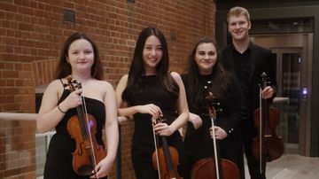 Four musicians in formal attire holding string instruments, standing in a brick-walled hallway, smiling at the camera.