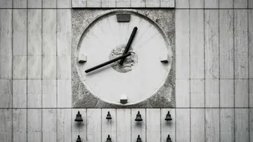 A large outdoor clock with black hands is mounted on a gray stone wall, showing the time as approximately 1:40, with a row of small bells positioned below it.