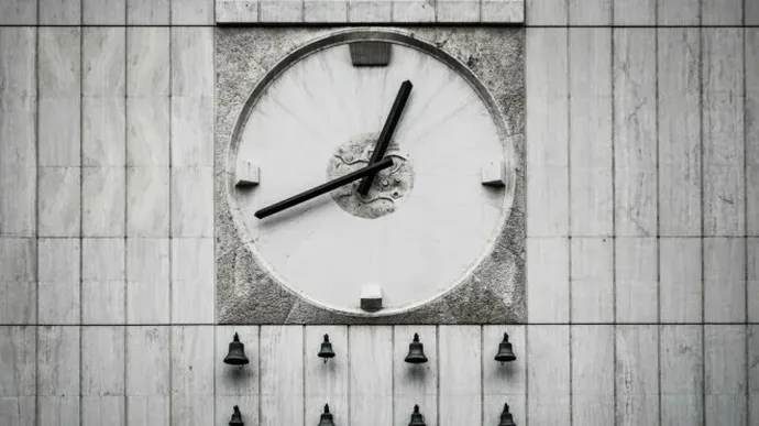A large outdoor clock with black hands is mounted on a gray stone wall, showing the time as approximately 1:40, with a row of small bells positioned below it.