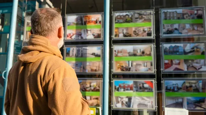 A person wearing a tan jacket looks at a display of property listings in an estate agent’s window, featuring photos of homes and green price banners.